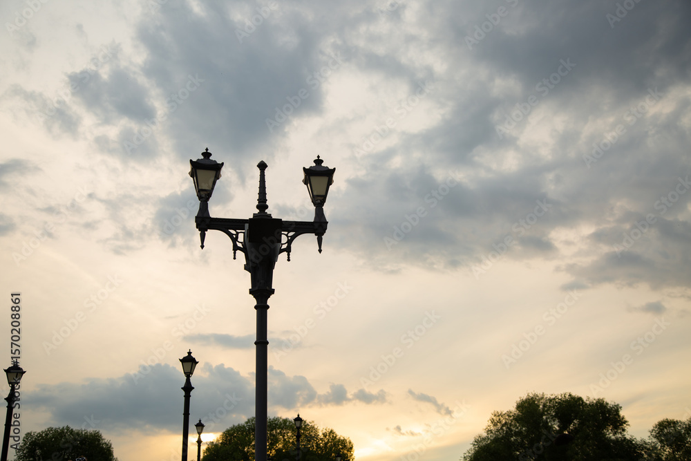 Street black iron design lamp in the park outdoors on summer green trees background under colorful sunset sky Street black iron design lamp in the park outdoors on summer green trees background under colorful sunset sky