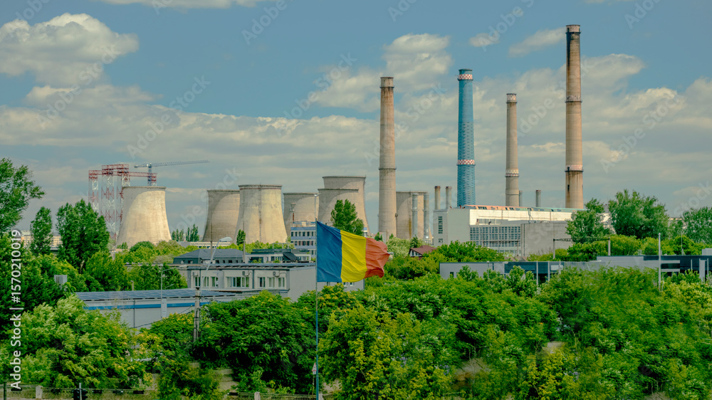 Obraz premium Romanian flag waving in front of a large industrial plant with multiple smokestacks and cooling towers on a partly cloudy day. 