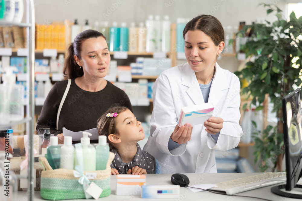 Obraz premium Polite young female pharmacist standing by computer showing medicament to mother with daughter in chemistry