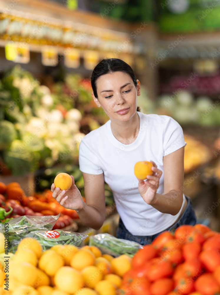 Obraz premium Woman near vegetable stand in store choose peach. Client view ripeness of fruit, take out fruits from showcase, collects grocery basket. Girl pull out ripest fruits from large pile of fruits.