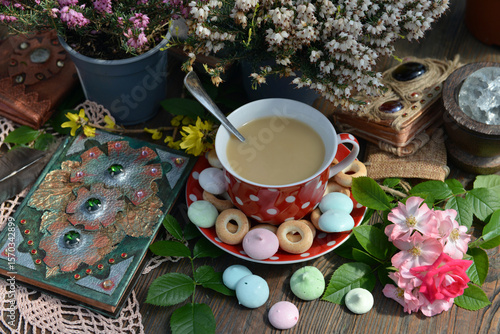 Cozy still life with cup of tea with milk, decorated book and sweets with flowers on the table. Romantic, vintage home and greeting card concept