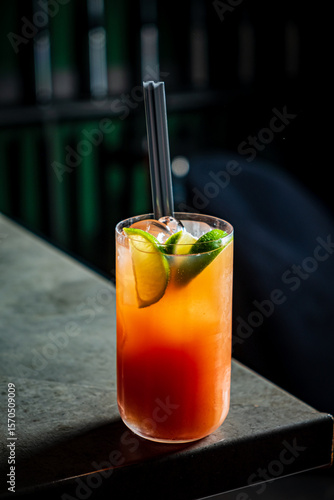 Brightly colored cocktail filled with lime wedges sits on a sleek bar countertop at night