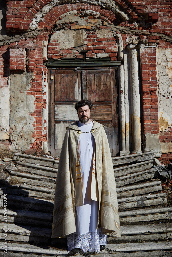 Naklejka premium Caucasian middle aged man standing in front of old church entrance wearing priest vestments, looking directly at camera with serious expression, stone steps and weathered brick wall visible
