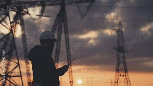An electrician on the background of high towers of power plants looks at the project for the development of an electrical structure, the expansion of the electrical voltage of volts in the wires.