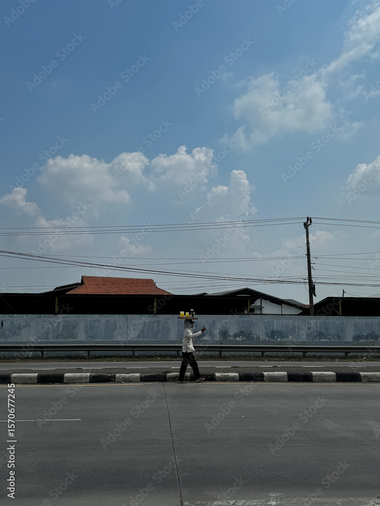 Fototapeta premium Demak-Indonesia, November 14, 2023: A street vendor balancing goods on his head, walking along a road on a sunny day.