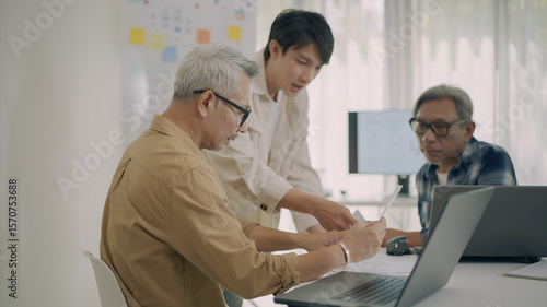 Team of Asian engineers discussing and reviewing software plan in office meeting. Concept of teamwork, innovation, and multigenerational learning in tech.