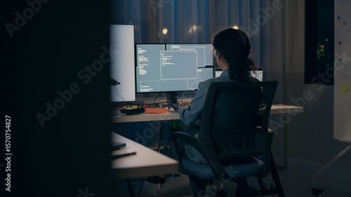 A dedicated female programmer seen from behind, intensely coding across multiple monitors in a dark office. Focuses on AI, machine learning, and software development.