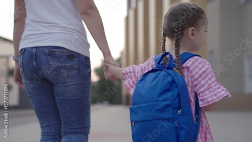little girl with backpack holds her mother by the hand and goes to school, kid with mom go to school in the school yard, child with school bag hurries to first lesson, preparatory preschool education.