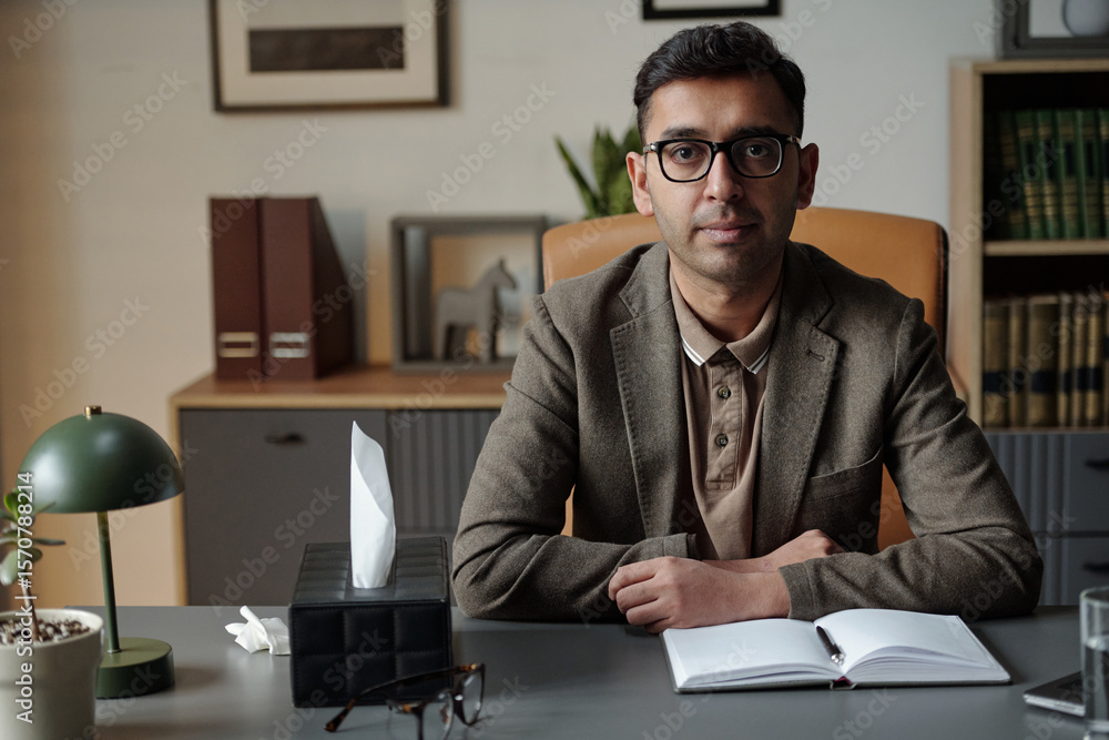 Fototapeta premium Portrait of man sitting at desk in office setting looking into camera, hands resting on open notebook, tissue box and eyeglasses on table, therapy context