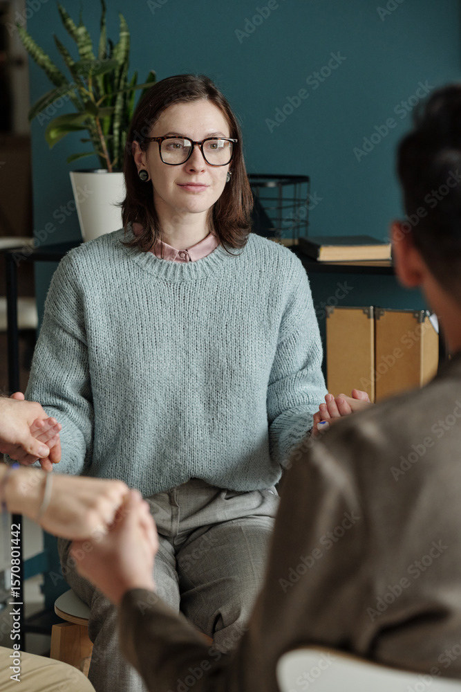 Fototapeta premium Woman sitting in therapy session holding hands with other participants, engaging in group support activity, looking forward with calm expression