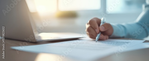 The hand writing on a document beside a laptop in a bright office setting.