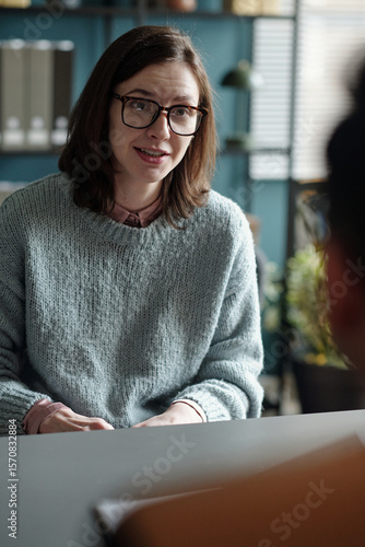 Woman sitting across from unseen individual engaging in therapy session, maintaining attentive expression, hands resting on table, bookshelf in background