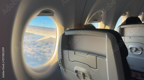 View on the fragment of the airplane cabin with passengers during the flight and a plane porthole through which you can see the wing of the plane and the blue sky with clouds in sunset.