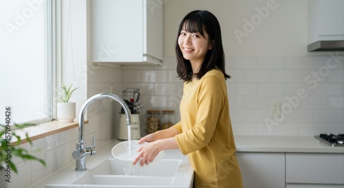 woman rinsing dishes in sink. Young Asian woman washing dishes in a bright kitchen with plants  