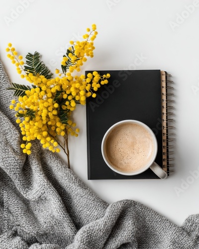 Flatlay of mimosa flowers, a grey knit blanket, a cup of coffee, and a black notebook on a white surface