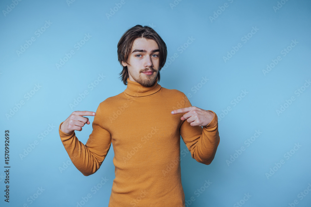 Naklejka premium Young man wearing a turtleneck sweater poses confidently against a solid blue background while pointing at himself during a casual moment