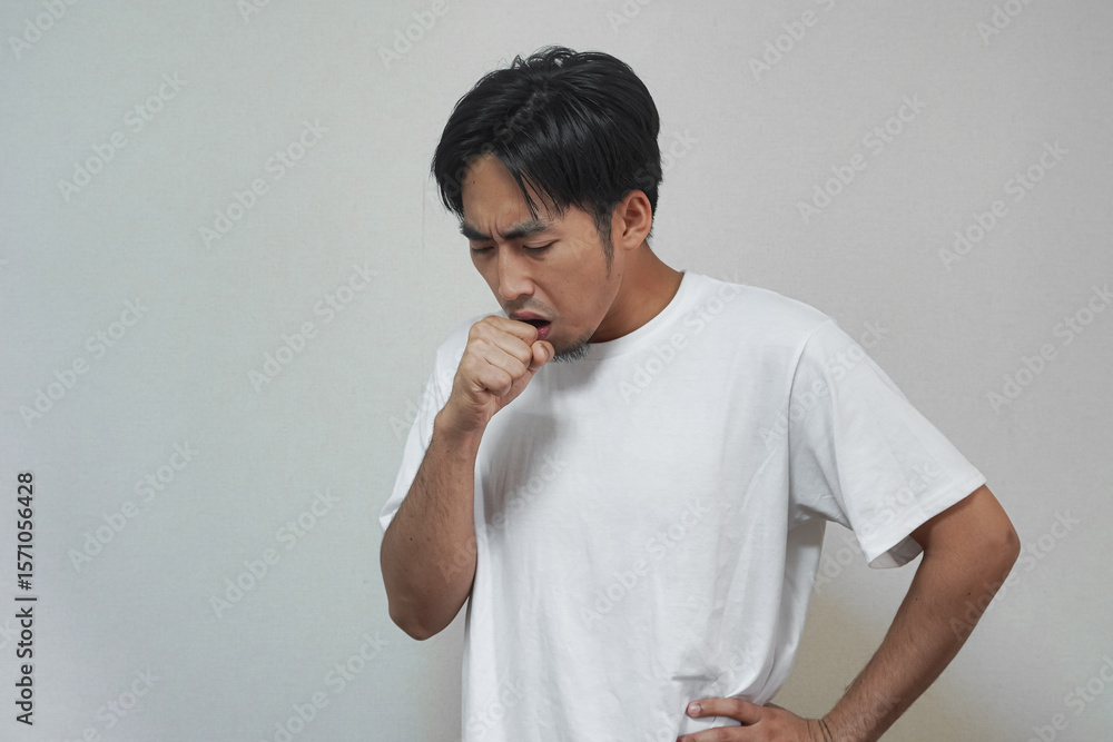 A young Asian man coughs, covering his mouth, with a hand on hip. A young Asian man coughs, covering his mouth, with a hand on hip.