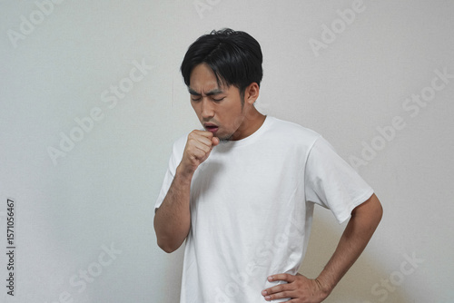 A man in a white t-shirt coughs into his hand, appearing unwell against a plain background.