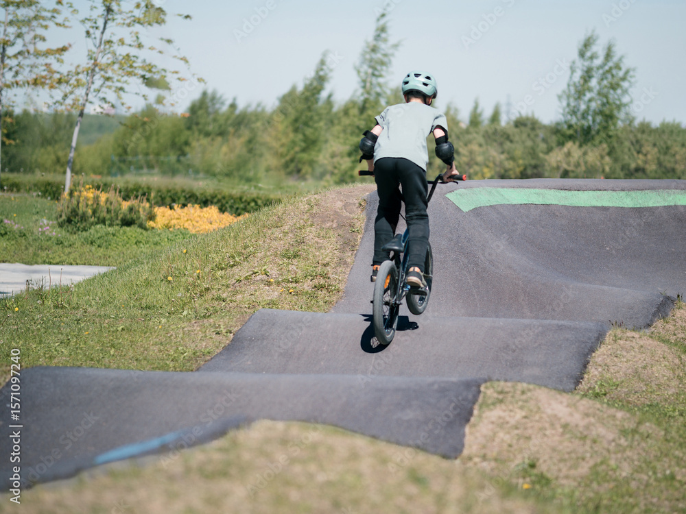 Fototapeta premium Child on bmx bike on pump track. Young male cyclist in helmet and protection rides bmx bike on asphalted bicycle race track. Boy BMX rider having fun and training on racing speed track. Extreme sport