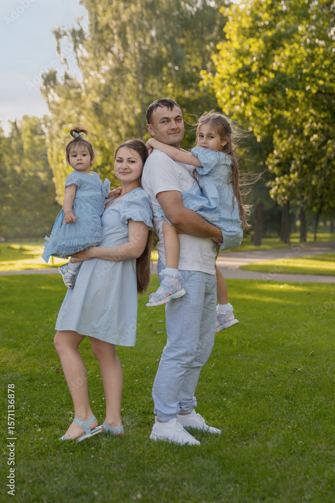 Obraz premium Mom and dad are standing back-to-back, holding their daughters in their arms. The family is dressed in blue, and everyone is looking at the camera. The concept of family, raising children, daughters,