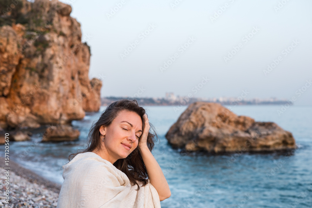 A young Caucasian woman with long brown hair smiles while sitting on a beach. She wears a light shawl and enjoys the serene ocean view with rocky cliffs in the background. A young Caucasian woman with long brown hair smiles while sitting on a beach. She wears a light shawl and enjoys the serene ocean view with rocky cliffs in the background.