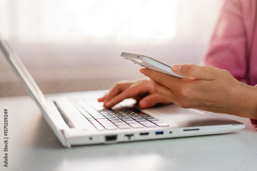 Fototapeta premium Hands type on a laptop keyboard while holding a smartphone, showcasing multitasking in a modern work environment.