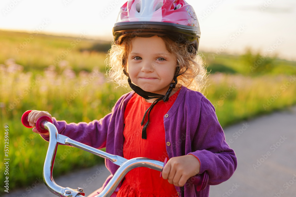Obraz premium A cute little girl in a pink helmet and a purple cardigan looks directly at the camera while holding her bicycle handlebars, with a sunlit grassy field in the background.