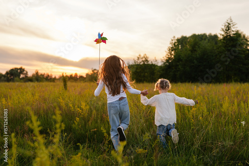 Quadro su tela A joyful older sister with a pinwheel and her younger sister run hand-in-hand through a tall, sunlit field at sunset