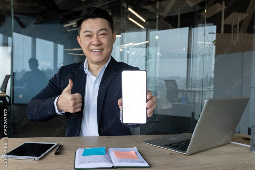 Portrait of a young Asian man in a suit sitting in an office at a desk, showing a white blank phone mockup to the camera and making a super sign with his finger Portrait of a young Asian man in a suit sitting in an office at a desk, showing a white blank phone mockup to the camera and making a super sign with his finger
