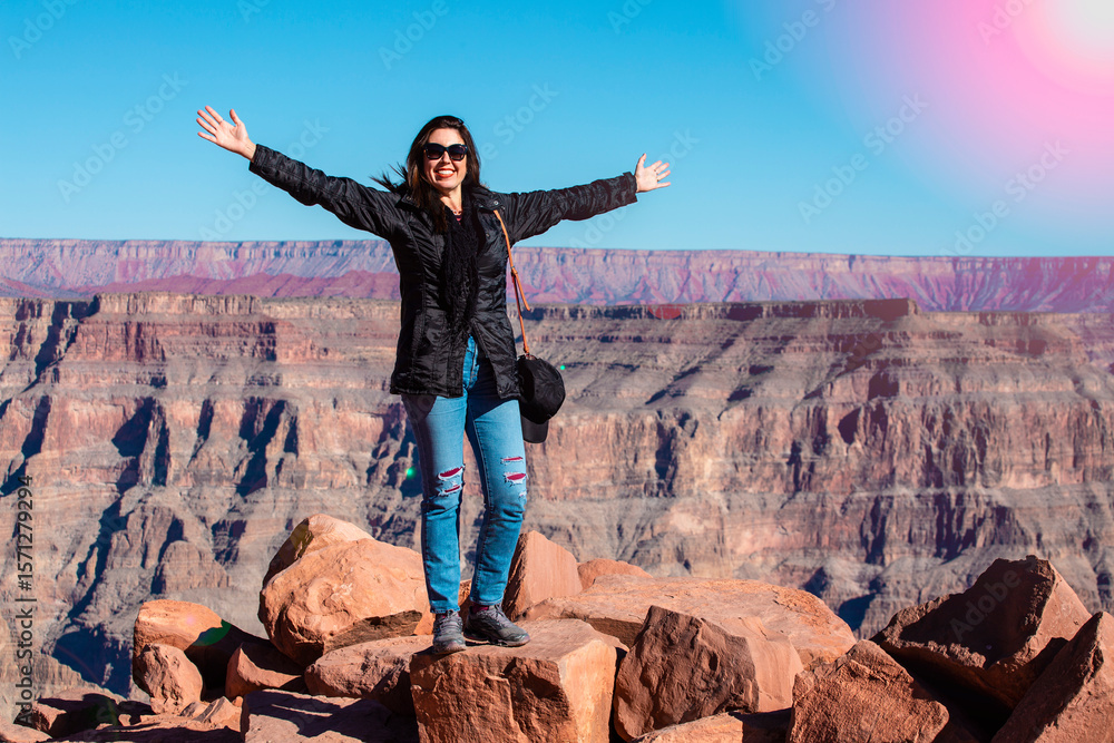 Obraz premium Happy woman standing on rocks with arms wide open, enjoying the scenic view of a vast canyon under a clear blue sky. Adventure, freedom, and travel inspiration in nature.