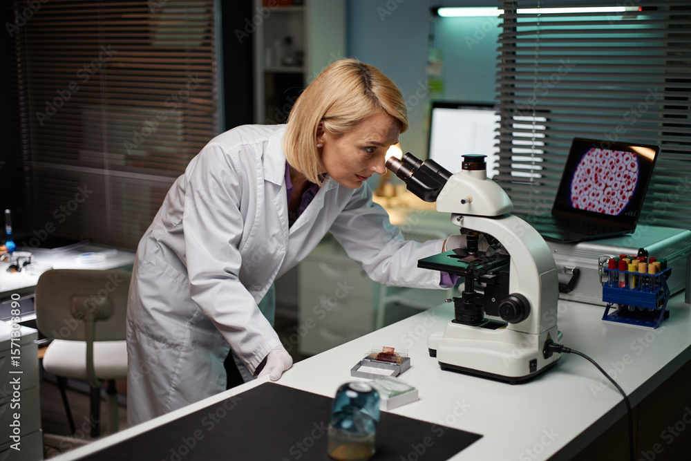 Middle aged Caucasian woman examining sample through microscope in scientific laboratory, focusing on research process with laboratory equipment and computer displaying cell images nearby Middle aged Caucasian woman examining sample through microscope in scientific laboratory, focusing on research process with laboratory equipment and computer displaying cell images nearby
