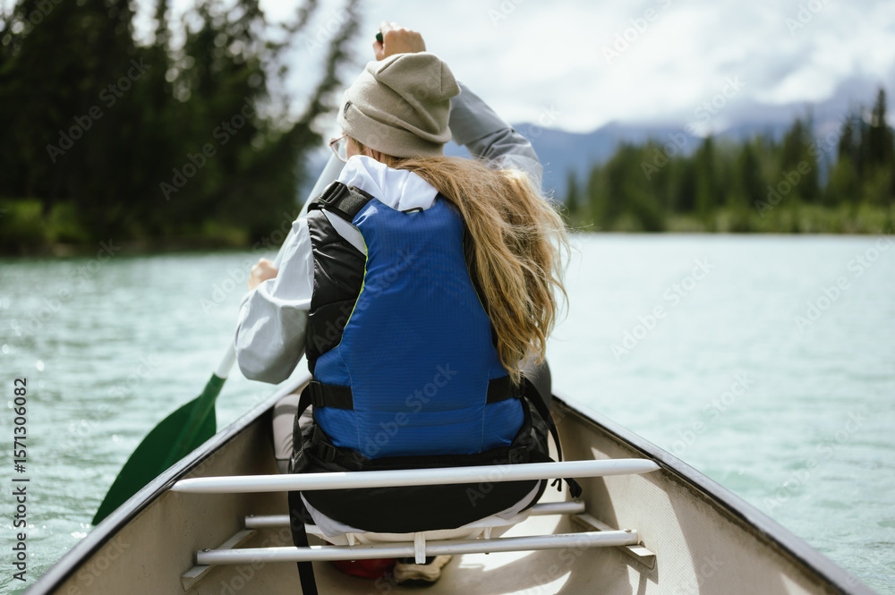 Back view of a young woman paddling a canoe across a clear, turquoise lake surrounded by pine trees and distant mountains with copy space Back view of a young woman paddling a canoe across a clear, turquoise lake surrounded by pine trees and distant mountains with copy space
