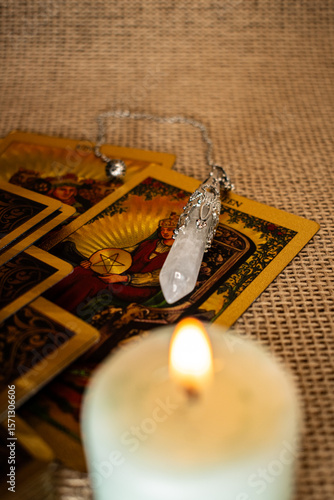 Tarot cards with vivid illustrations lie on burlap, with a quartz pendulum and a lit candle. A mystical setup evoking divination, fortune-telling, and spiritual ritual themes.
