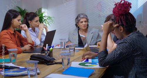 Indian young adult woman staff sitting around desk table with old aged boss ceo lady talk share bad news at job place upset female leader speak about work loss to girl group team office board room