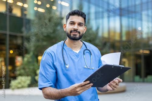 A medical professional, likely a doctor or nurse, stands outside a building, holding a clipboard and looking at the camera.