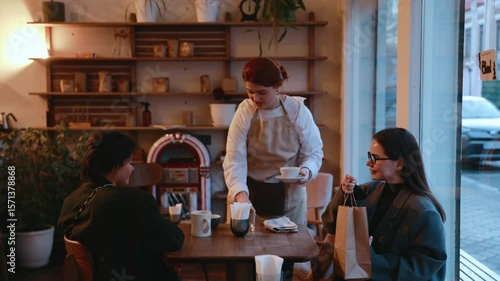 A female employee puts down a towel and takes away cups from a table from which two female customers are getting up