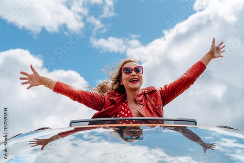 Joyful woman enjoying a road trip with arms raised in the air under a blue sky with fluffy clouds during daytime summer escapade