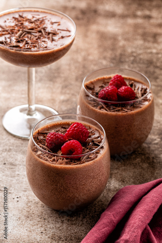 Homemade chocolate mousse decorated with raspberries and dark chocolate sprinkles in portion glasses. Brown table surface. Vertical image, selective focus.