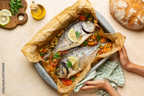 Hands holding cooking tray with baked Dorado fish or sea bream with tomatoes and olives, fresh bread, olive oil, lemon. Mediterranean food. Healthy dinner, top view.