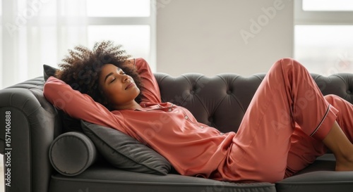 Relaxing Woman in Pink Pajamas on Sofa. A woman in cozy pink pajamas reclining on a stylish gray sofa in a sunlit room, capturing a moment of relaxation and comfort in an elegant