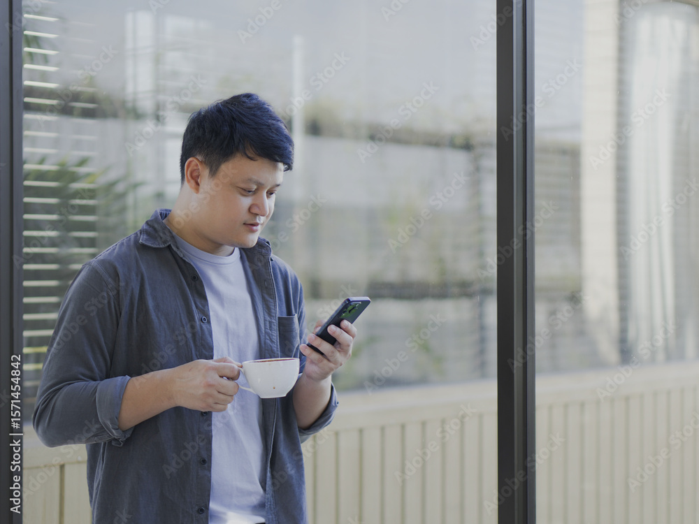 Fototapeta premium A young Asian man in casual clothes is holding a coffee cup and looking at his mobile phone.