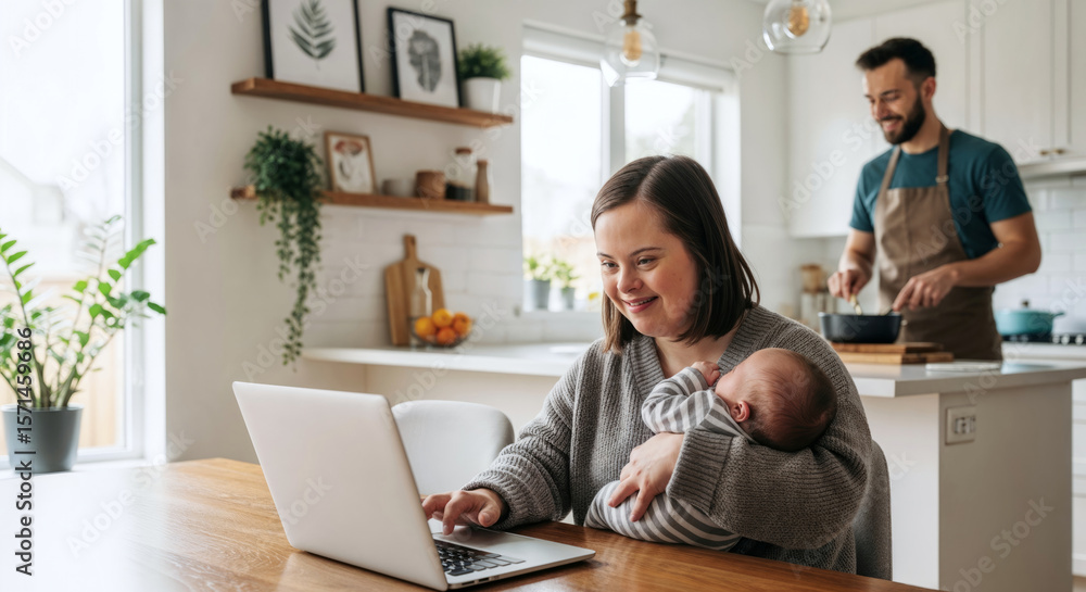 Fototapeta premium Young mother with down syndrome holding her sleeping newborn baby while working remotely from home on a laptop with her partner cooking in the background. Inclusive and supportive family life