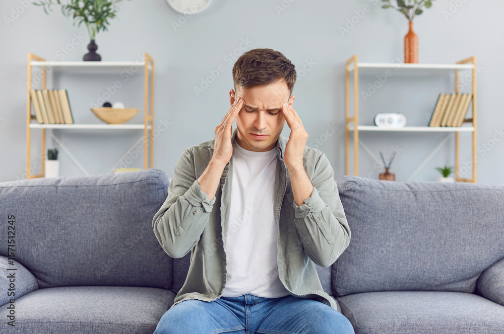 Fototapeta premium A young man sits on the sofa at home alone and touches his head, close-up. Migraine and headache concept. Reaction to severe stress, emotional burnout. Depression and loneliness