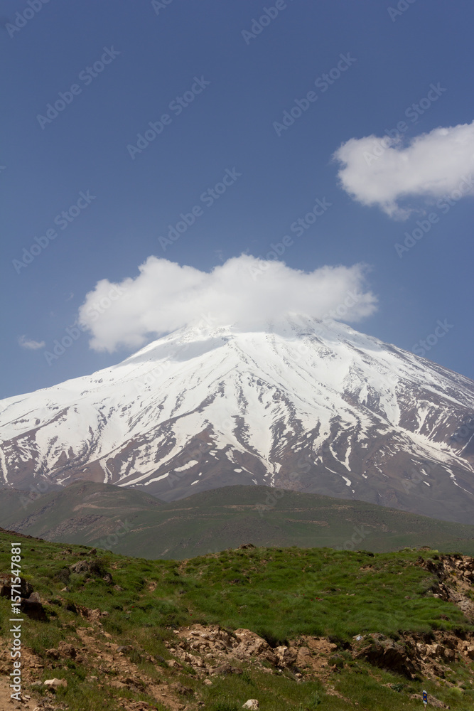 Obraz premium Majestic Mount Damavand rises above the Alborz range, crowned with snow and surrounded by lush hills and rugged terrain under a clear sky.