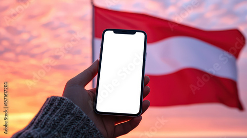 Welcome to Austria. Travel and tourism in Europe. A mockup of a cell phone with an empty white screen in a woman's hand against the background of the Austrian flag fluttering in the wind at sunset