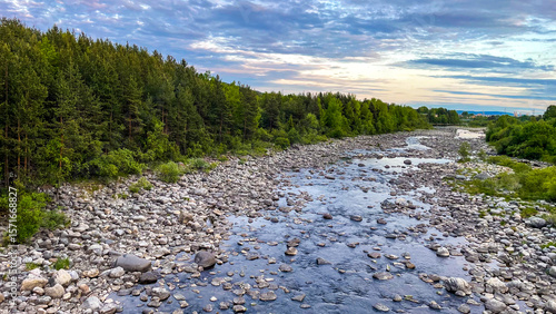 Fotografie Scenic view of a shallow stony river flowing through a green forest under a dramatic evening sky