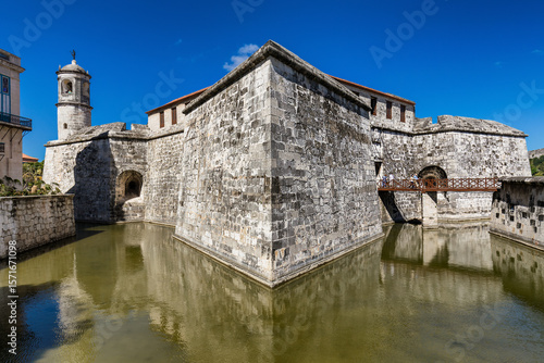 View of a formidable stone fortress reflecting in a still moat under a vibrant blue sky, a testament to history and strength, Havana, Havana, Cuba.