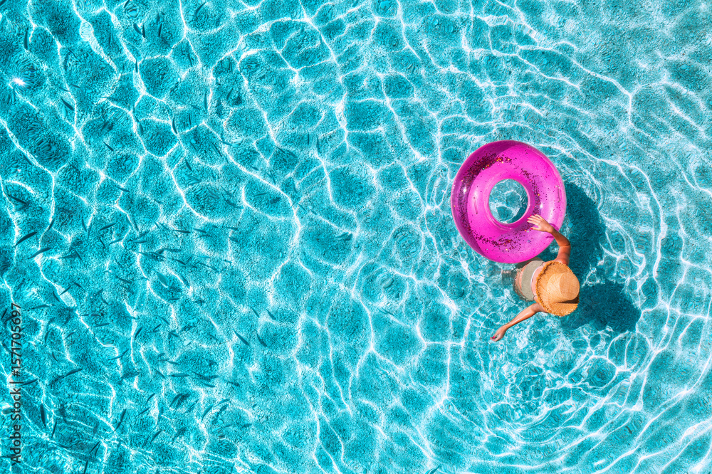 Naklejka premium Aerial view of a young woman in hat swimming with pink swim ring in blue sea with school of fish at sunset in summer. Tropical landscape with girl, clear water, sandy beach in Greece. Top view. Travel