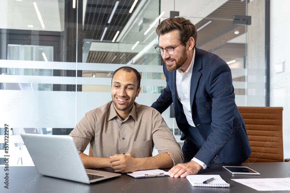 Obraz premium Two colleagues are collaborating on a project, one is seated at a desk with a laptop, while the other leans in to offer support.