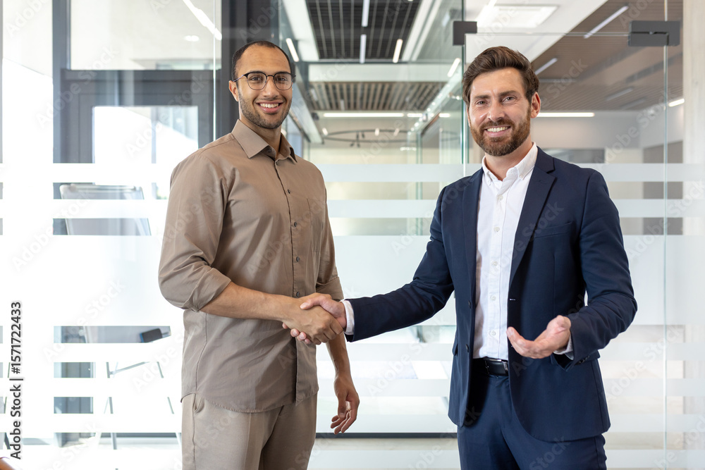 Fototapeta premium Two businessmen shake hands, smiling, in a modern office setting with glass walls. A gesture of agreement or partnership.